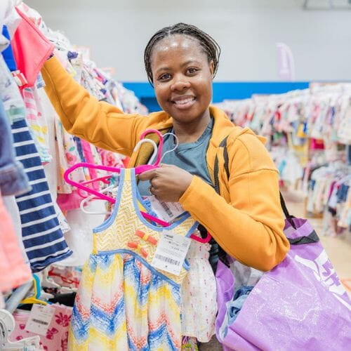 A mom with a large JBF shopping bag on her shoulder stands beside her husband who wears their toddler at a JBF sale.